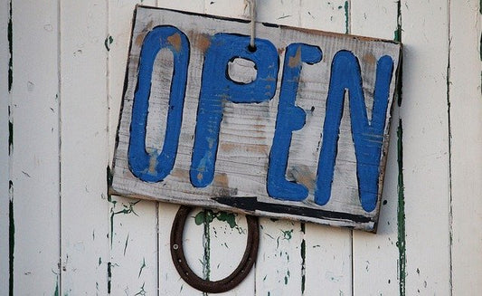 A wooden sign with the words open written in blue hanging on a white wooden wall.