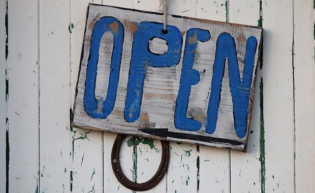 A wooden sign with the words open written in blue hanging on a white wooden wall.