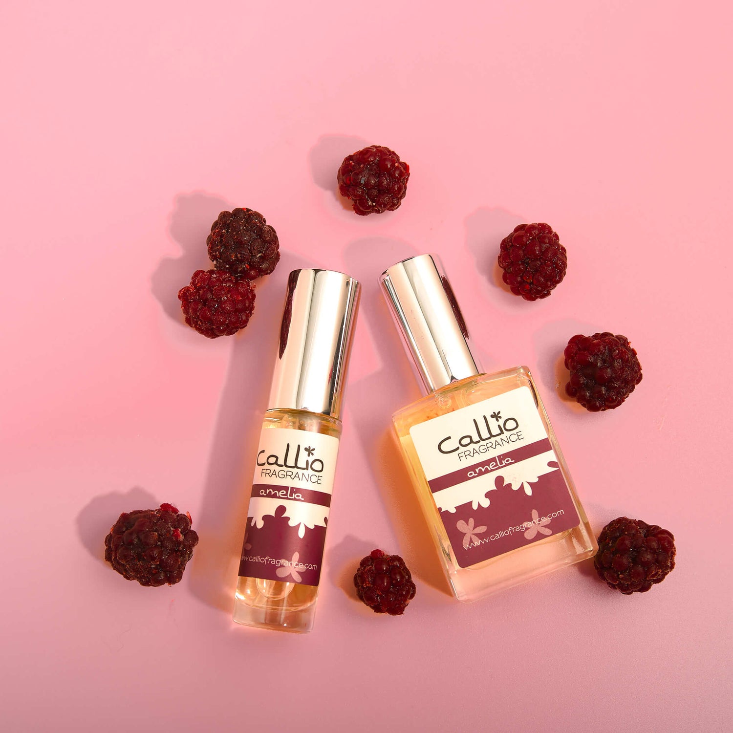 Travel-size Amelia and one-ounce bottle of Amelia on a pink background with blackberries next to the bottles. 